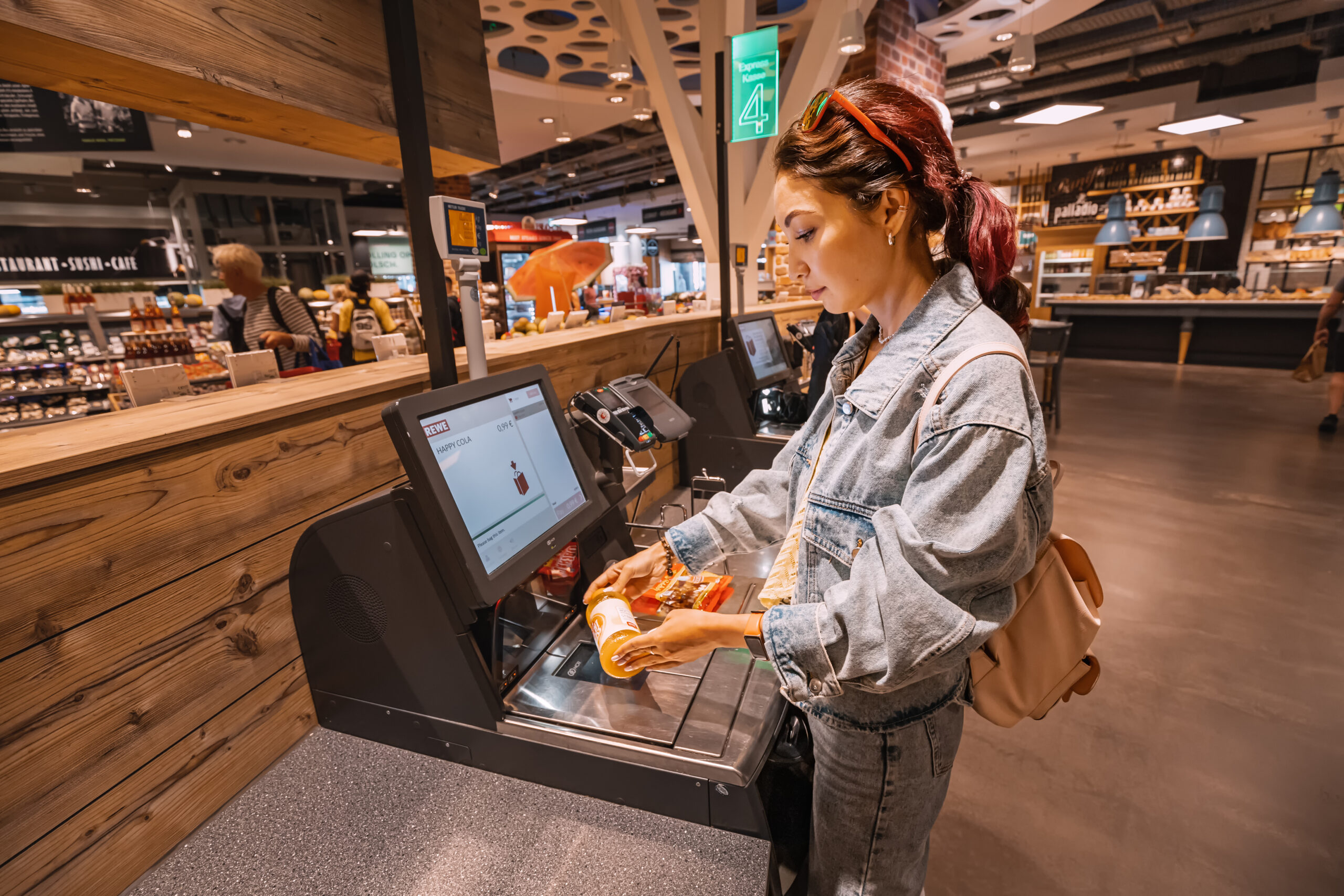 30 July 2022, Cologne, Germany: girl customer scanns and pays for bottle of juce at an automated self-service checkout terminal in Rewe supermarket 30 July 2022, Cologne, Germany: girl customer scanns and pays for bottle of juce at an automated self-service checkout terminal in Rewe supermarket